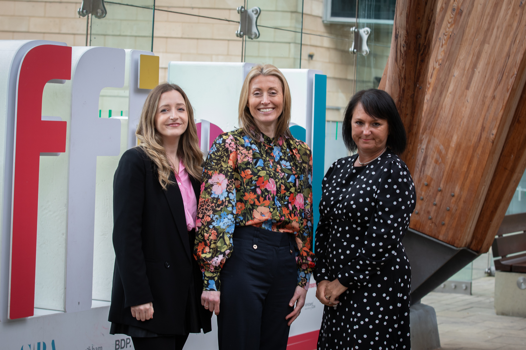 Three people standing in front of a large colorful sign that appears to spell out ‘Festival’ in an outdoor urban setting. The sign features bold letters in white, red, and teal. Behind them is a modern building with light stone walls and glass panels, and part of a large wooden architectural structure is visible on the right.
