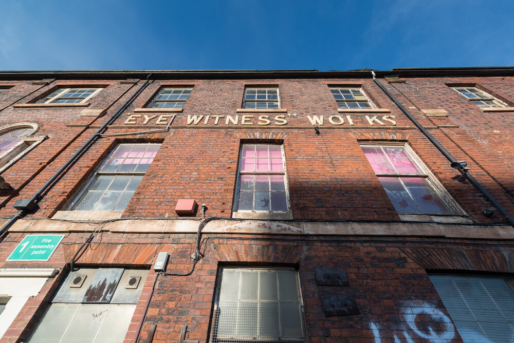A three-story brick building with large windows, some featuring pink-tinted glass. A faded sign reads 'EYE WITNESS WORKS' across the facade. A green 'Fire assembly point' sign is visible on the left side. The sky is clear and blue.