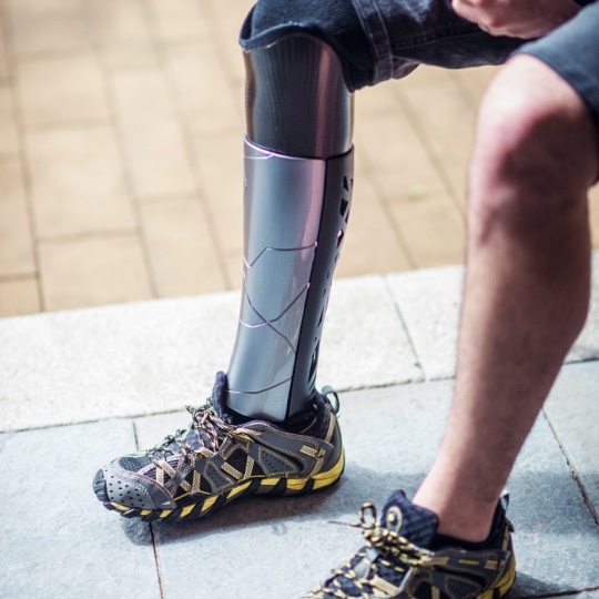 Close-up of a person seated outdoors wearing a sleek, metallic prosthetic leg paired with a rugged hiking shoe. The prosthetic has a modern, angular design with a smooth finish, and the person’s other leg is bare, wearing a matching shoe. The background shows a paved surface and a stone edge.