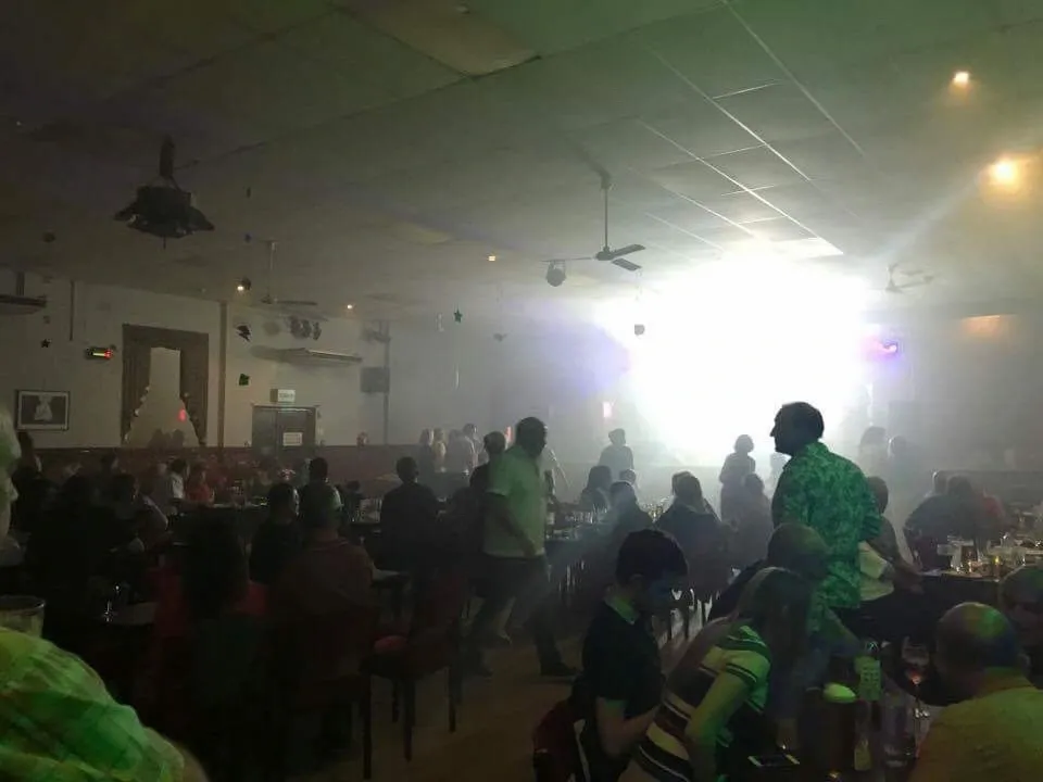 Interior of Crookes Social Club - A crowded indoor venue with people seated at tables and others standing, creating a lively social atmosphere. The room is dimly lit with a bright light source in the background, casting a hazy glow and adding a smoky effect. Ceiling fans and hanging lights are visible above, and drinks and glasses are placed on tables throughout the scene.