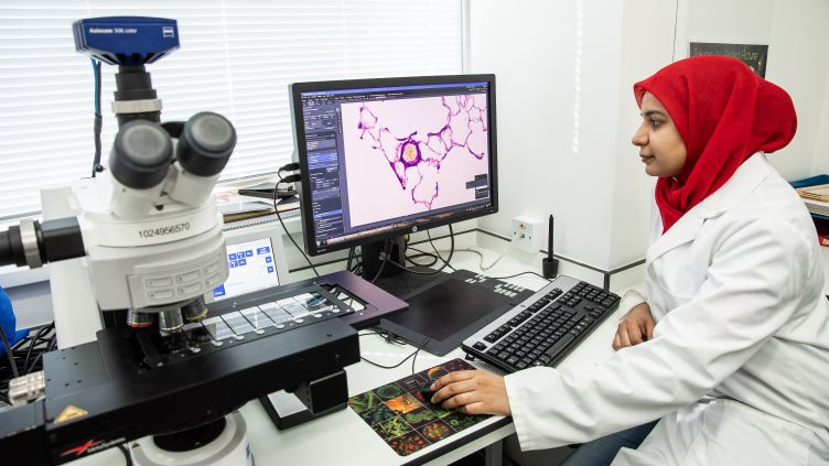 A woman in a white lab coat and a red headscarf sat at a desk in a laboratory using a computer.