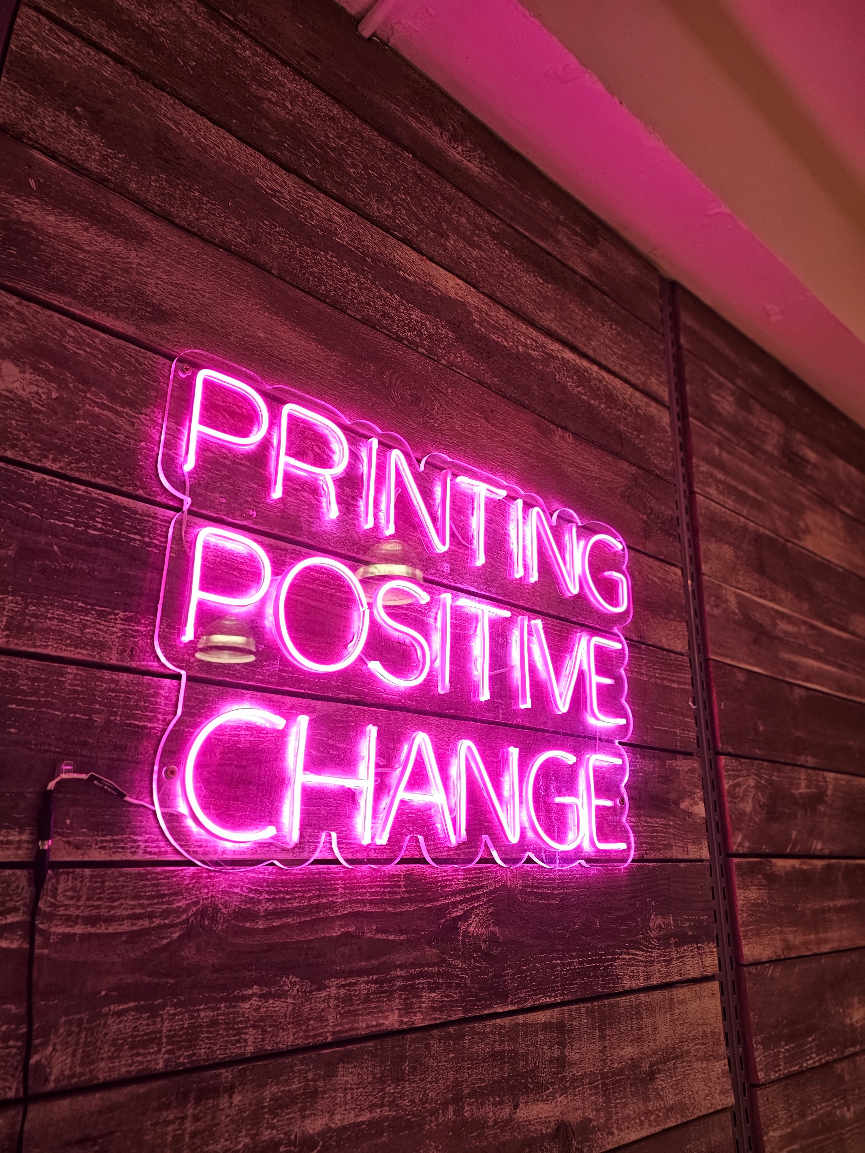 A pink neon sign that reads 'Printing Positive Change'.