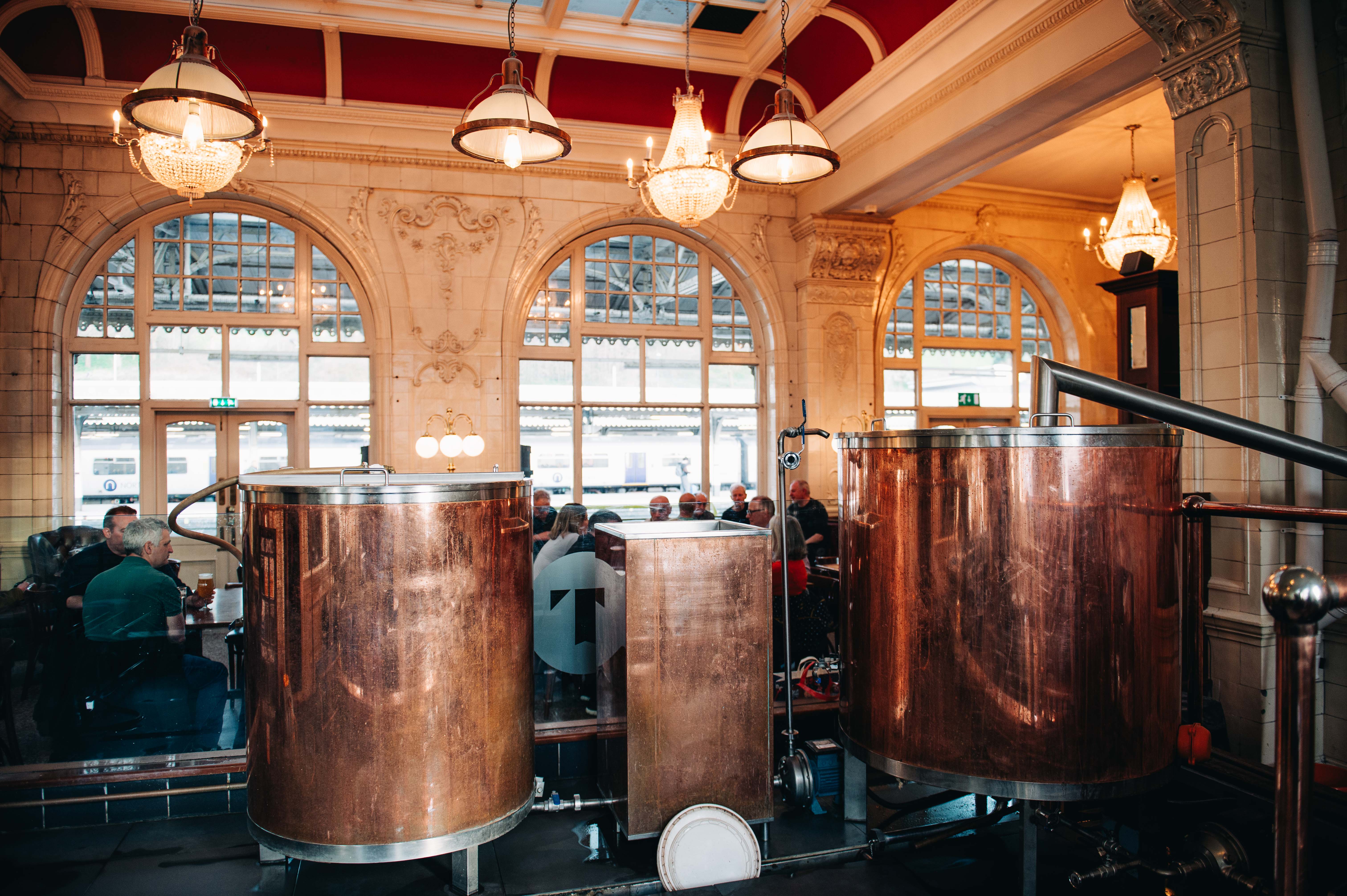 Sheffield Tap copper beer tanks. Groups of people are sat talking and drinking, with white chandeliers hanging from the ceiling, and floor to ceiling windows looking onto the station platforms.