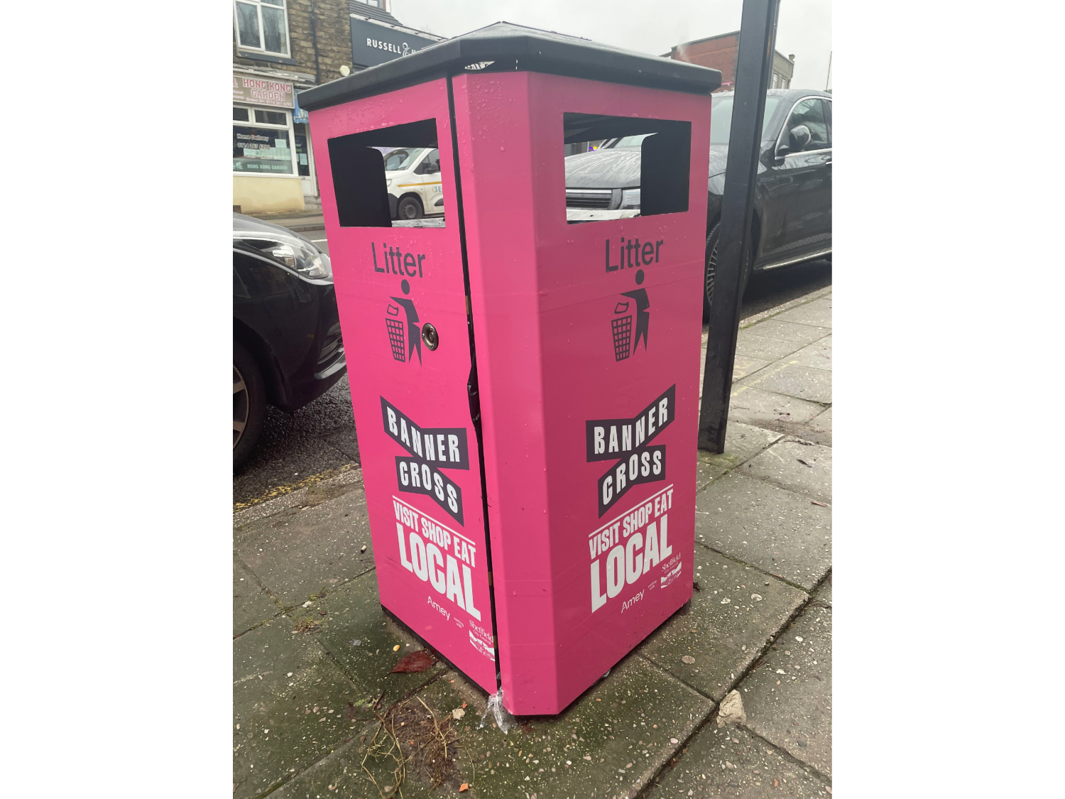 Bright pink litter bin on a pavement with text reading ‘Banner Cross’ and ‘Visit Shop Eat Local’. The bin has two openings at the top and is positioned near parked cars and a shopfront.