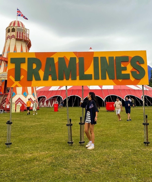 Joey, a third-year Festival and Entertainment Management student at Sheffield Hallam University, at Tramlines Festival in Sheffield. A large colorful sign reading “TRAMLINES” stands on a grassy field at a festival site. Behind the sign is a red and white striped big top tent and a tall helter-skelter slide with a Union Jack flag on top. Several people are walking around the area under a cloudy sky.