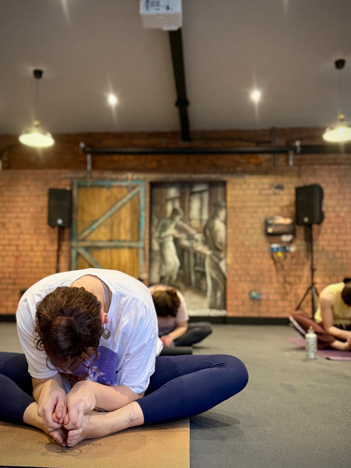 Indoor yoga class in a brick-walled studio with industrial-style lighting and a mural in the background. Several people are seated on yoga mats performing a forward bend stretch, holding their feet with both hands. A water bottle is visible on the floor near one participant.