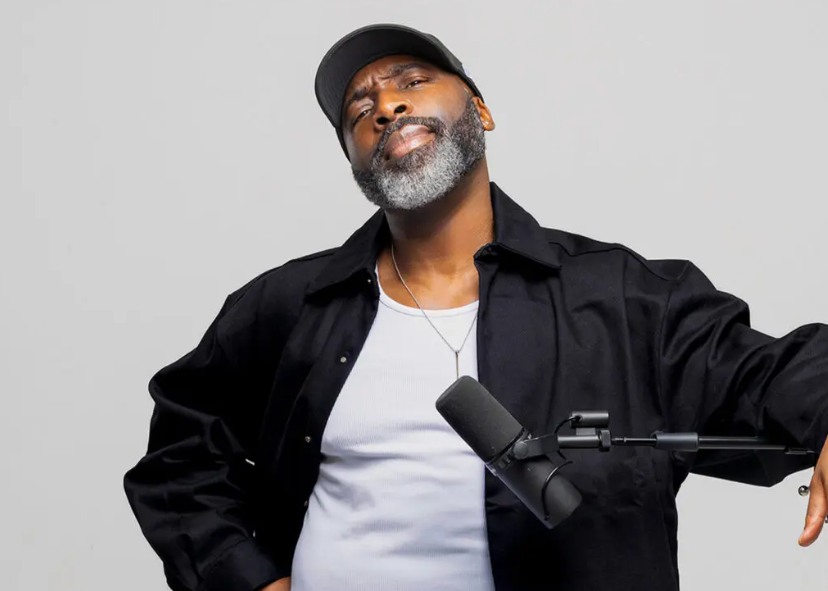 Person wearing a black jacket over a white shirt and a necklace, standing in front of a plain light gray background. The person is holding one arm out to the side and is positioned near a black microphone mounted on a boom arm. A dark cap is worn on the head.