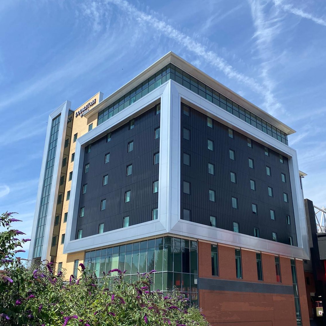 Modern multi-story hotel building with a distinctive design featuring a large white rectangular frame around dark panels with small windows. The structure includes glass sections at the lower level and a tan-colored tower on the left with the DoubleTree by Hilton logo. The sky above is bright blue with streaks of white clouds, and greenery is visible in the foreground.