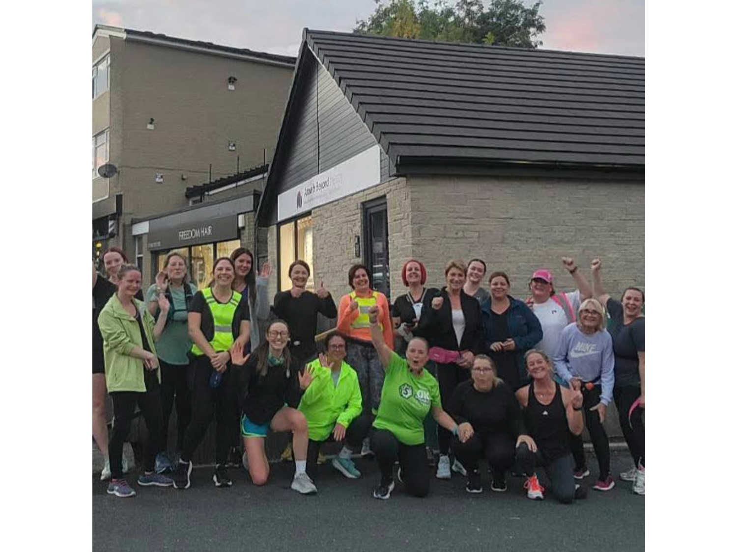 Group of around 20 people standing outside a building with a sign reading "Sports & Beyond Fitness." They are smiling and waving at the camera, dressed in a mix of athletic and casual clothing. Urban surroundings with other buildings in the background.
