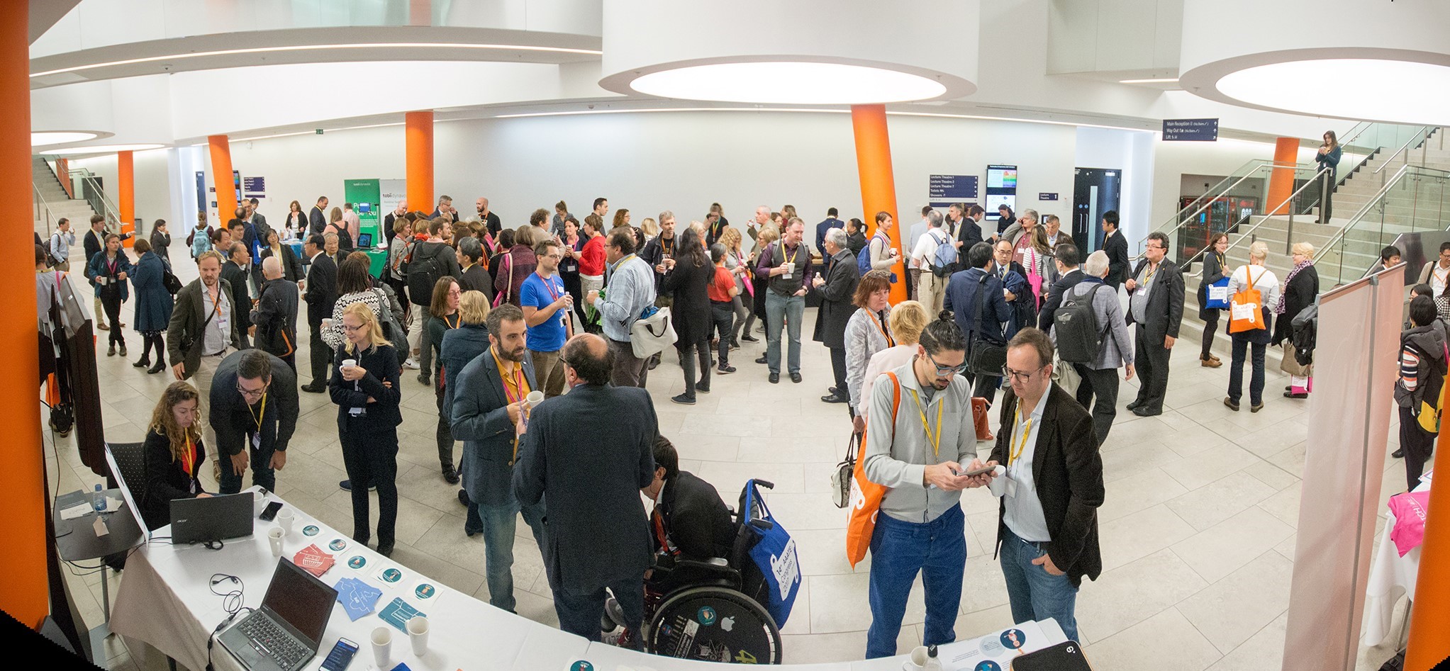 Large networking event in a bright, modern conference space with white walls and orange columns. Dozens of attendees are gathered in groups, talking and holding event bags. A registration table with laptops and promotional materials is visible in the foreground, and a staircase with glass railings is in the background.
