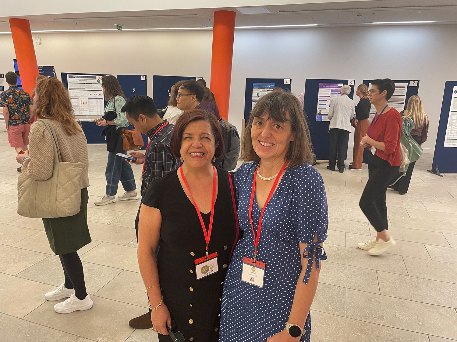 Conference Ambassadors collaborate to host ISA Conference. Two individuals wearing conference badges and red lanyards stand in the foreground of an indoor event space. Behind them, several people are engaged in conversation and viewing research posters displayed on dark blue boards. The setting appears to be a professional or academic conference with bright lighting and orange structural columns.