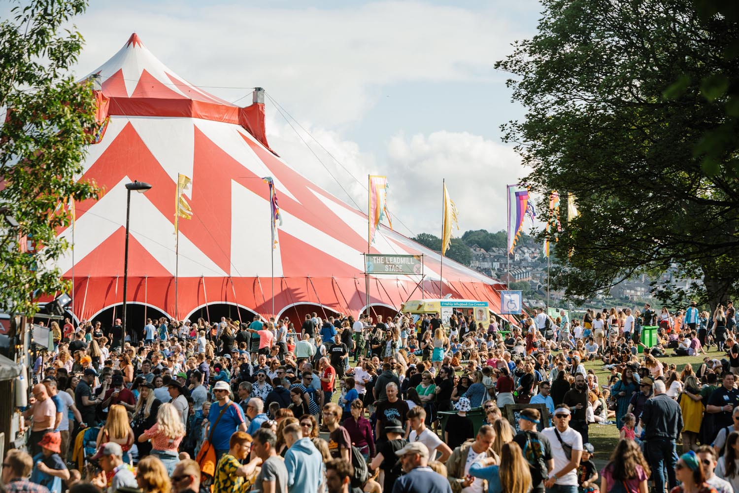 A crowd of people at the 2019 Tramlines Festival at Hillsborough Park in Sheffield. In the centre of the picture is a huge red and white circus-style tent.