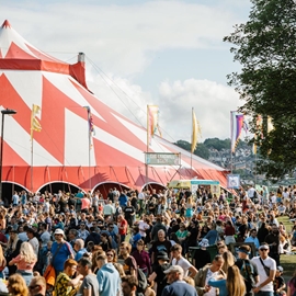 A crowd of people at the 2019 Tramlines Festival at Hillsborough Park in Sheffield. In the centre of the picture is a huge red and white circus-style tent.