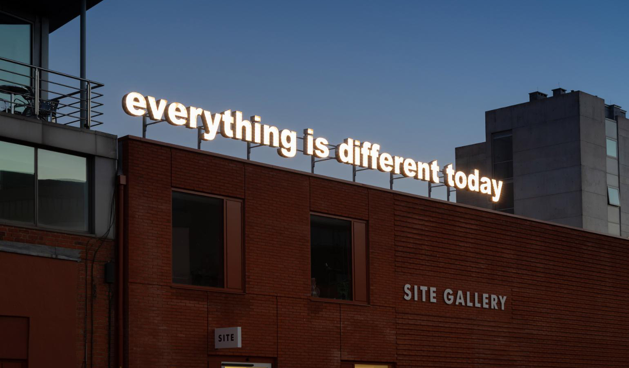 A red brick building. On the side is a sign that reads 'Site Gallery'. On the roof is an illuminated sign, made up of individual letters, that reads 'everything is different today'.