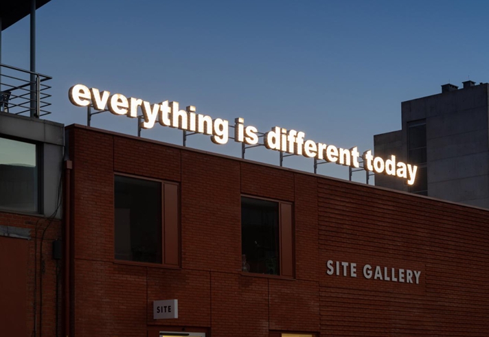A red brick building. On the side is a sign that reads 'Site Gallery'. On the roof is an illuminated sign, made up of individual letters, that reads 'everything is different today'.