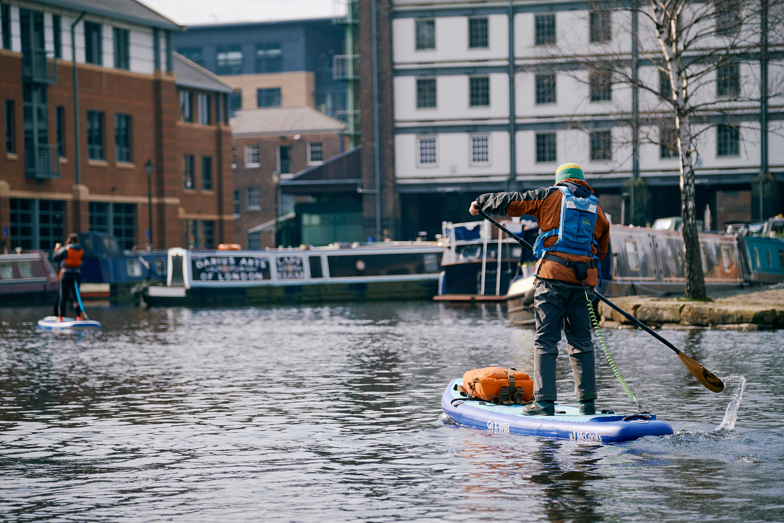 Two people are paddle boarding on the water at Victoria Quays in Sheffield city centre. There are several long boats moored around the edges of the water.