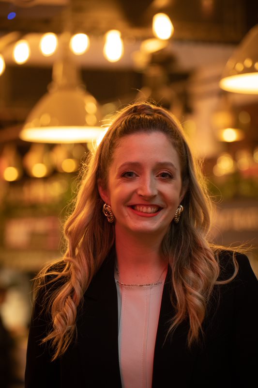 Person with long wavy hair wearing a light-colored top and a dark jacket, standing indoors under warm ambient lighting with hanging lamps and blurred background decor.