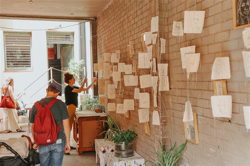 Indoor passageway with brick walls displaying numerous small sketches and drawings hung on strings. Several people are viewing the artwork, including one person with a red backpack walking toward the display and another adjusting pieces on the wall. Potted plants and a wooden table are placed along the corridor.