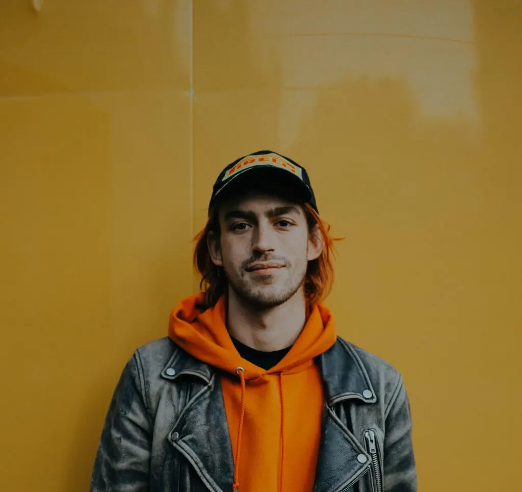 Person wearing an orange hoodie layered under a dark gray leather jacket and a black cap with a colourful patch, standing against a solid yellow background.