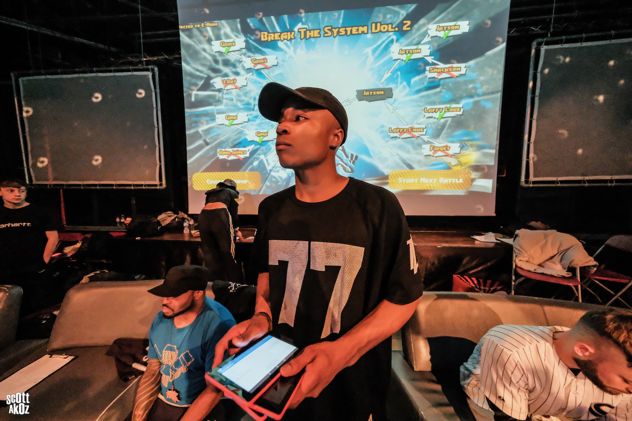 A person wearing a black T-shirt with the number “77” printed on the front is standing indoors holding a tablet device. Behind them, a large screen displays colorful graphics with the text “Break the System Vol. 2” and various game-related elements. Other people are seated on couches in the background, and the setting appears to be a gaming or esports event.