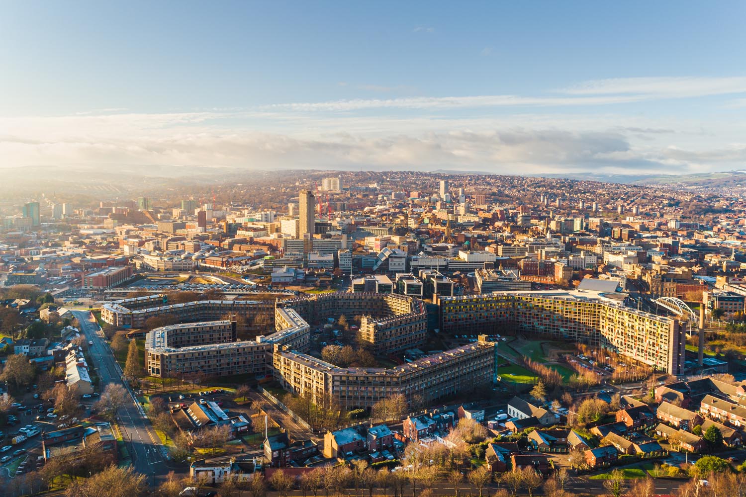 Aerial view of Sheffield cityscape under clear skies, showing a mix of modern and historic buildings. Prominent curved housing blocks are in the foreground, with the city center and tall structures in the distance. Hills and scattered clouds frame the horizon.