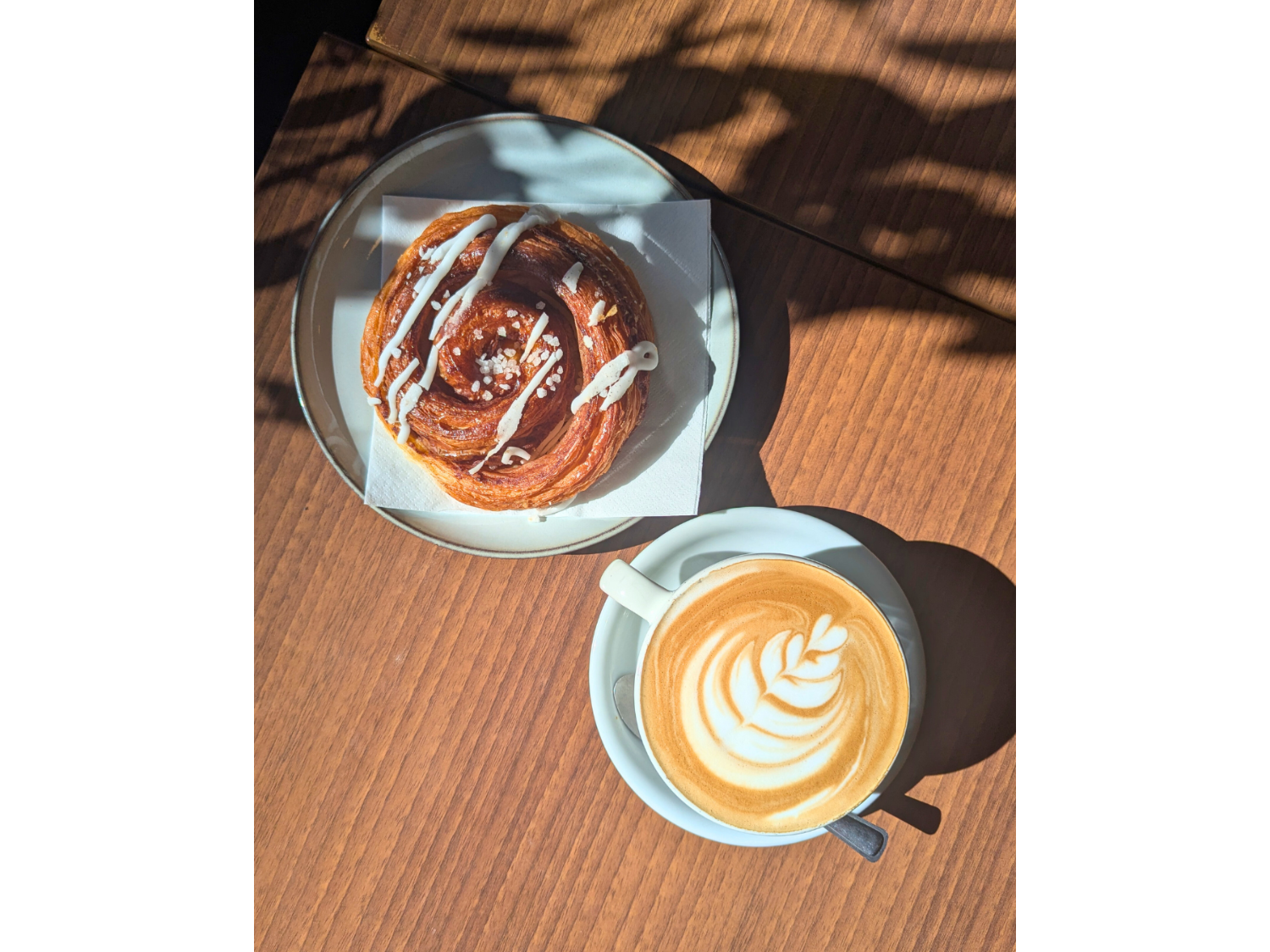 A wooden table, in the sunshine, with a danish pastry and a cappuccino. 