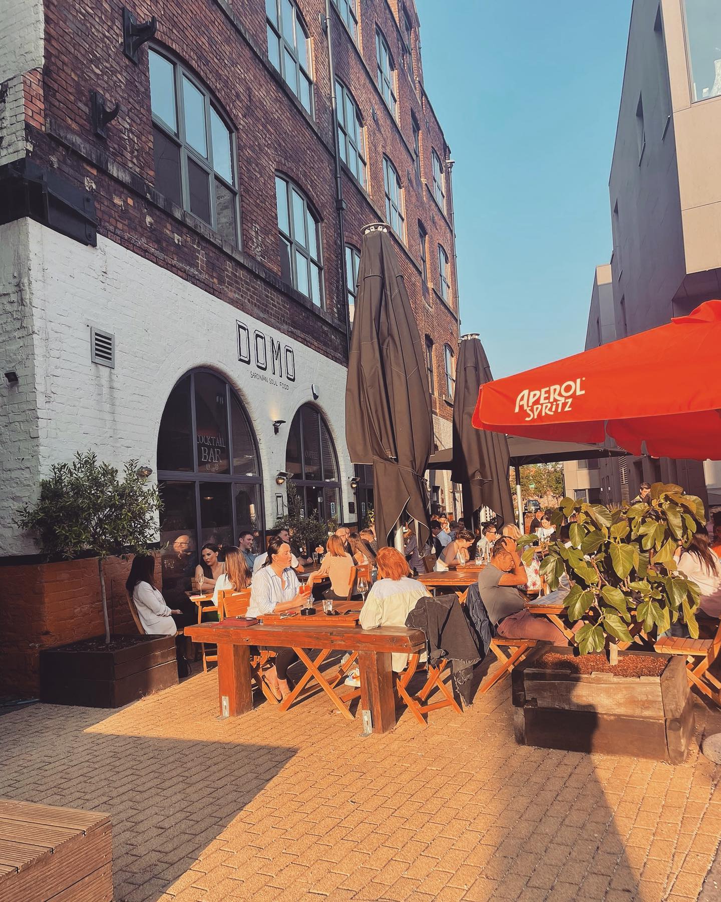 Outdoor seating area of a restaurant called “DOMO” located in a brick building with large arched windows. Several wooden tables and benches are arranged along the walkway, with people dining and socializing under tall black umbrellas. A bright red Aperol Spritz-branded umbrella adds colour to the scene, and potted plants provide greenery. The setting is sunny, creating a warm and inviting atmosphere.