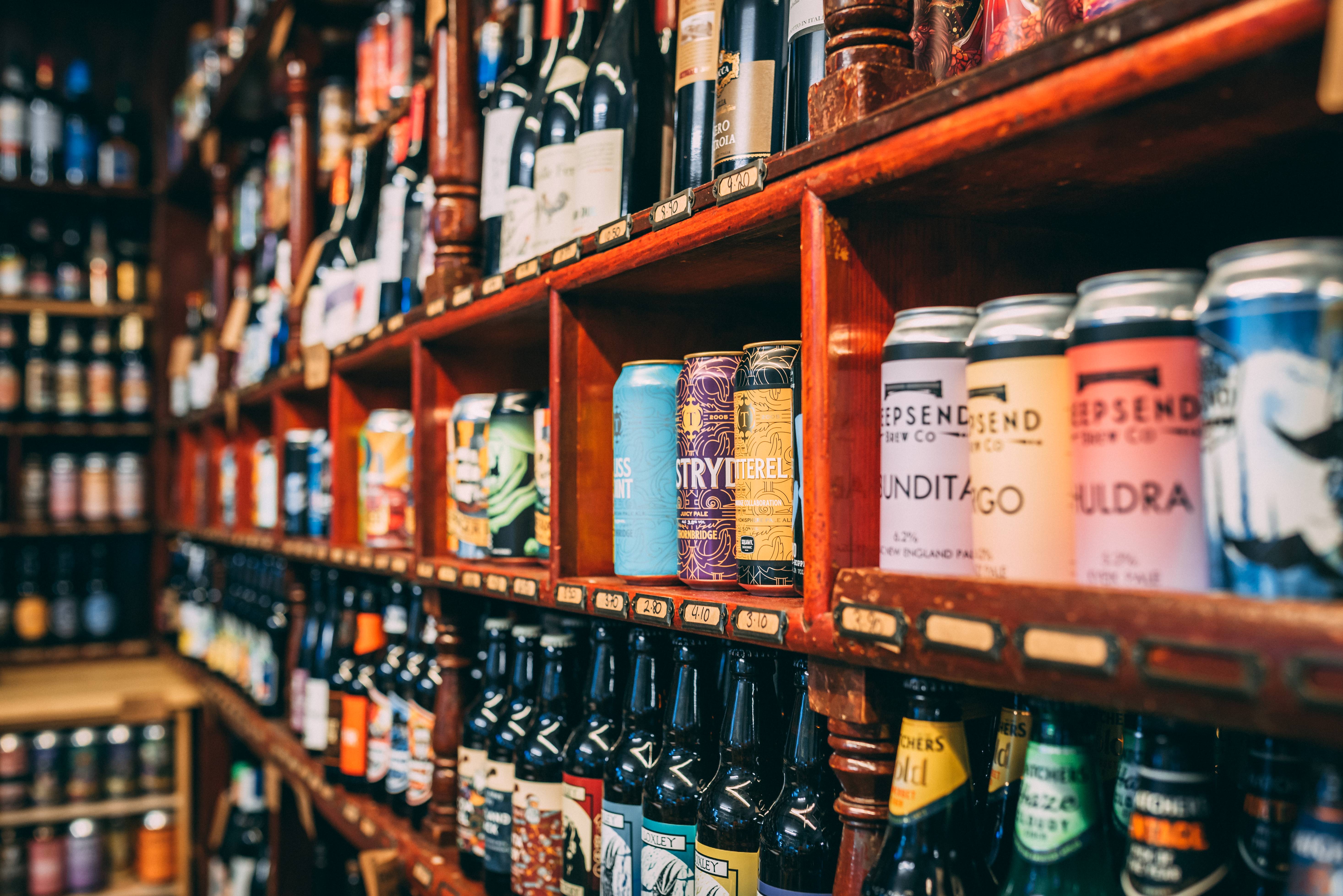 A store shelf filled with a wide variety of alcoholic beverages, including colorful cans and neatly arranged bottles of beer. Additional shelves in the background display more drink options, suggesting a large selection.