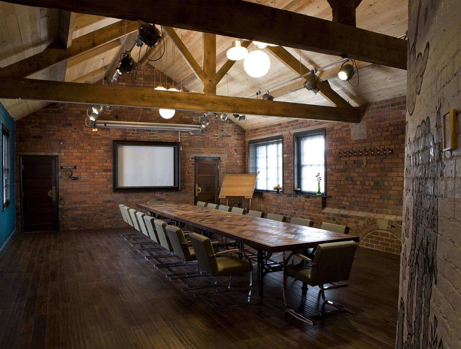 Rustic-style meeting room with exposed brick walls, wooden beams, and a long rectangular wooden table surrounded by chairs. The room features a projector screen on the far wall, two large windows, and a flip chart. Ceiling lights and track lighting provide illumination.