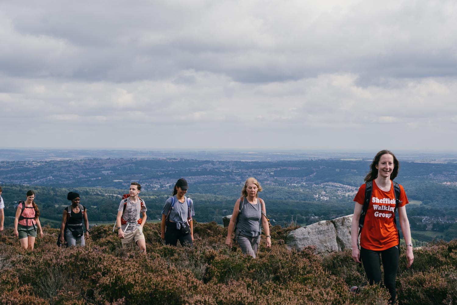A group of hikers walking uphill through a heathland area with low shrubs and rocky terrain. The hikers are wearing casual outdoor clothing and backpacks. In the background, there is a wide view of a valley with scattered buildings and greenery under a cloudy sky. One person in the foreground is wearing a bright red T-shirt with visible text that reads “Sheffield Walking Group