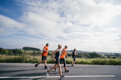 Four people running along a road, out in the country side. They are all wearing running gear.