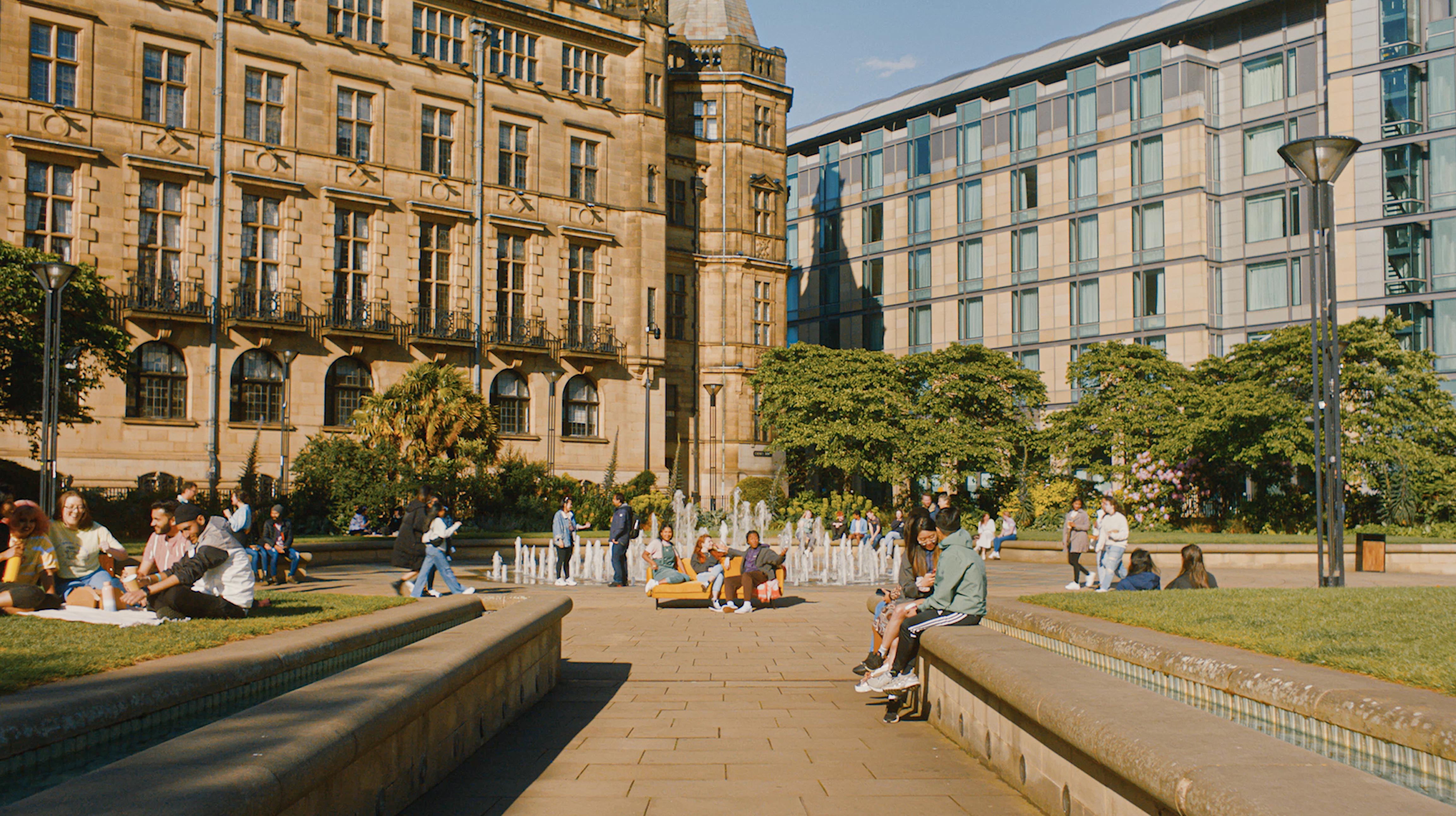 A sunny urban square with people sitting and relaxing near fountains and on benches. The scene features a large historic stone building with ornate architecture on the left and a modern glass-fronted building on the right. Trees and landscaped greenery add contrast to the paved area, and the sky is clear and bright. In front of the fountain are three people sat on a yellow couch.