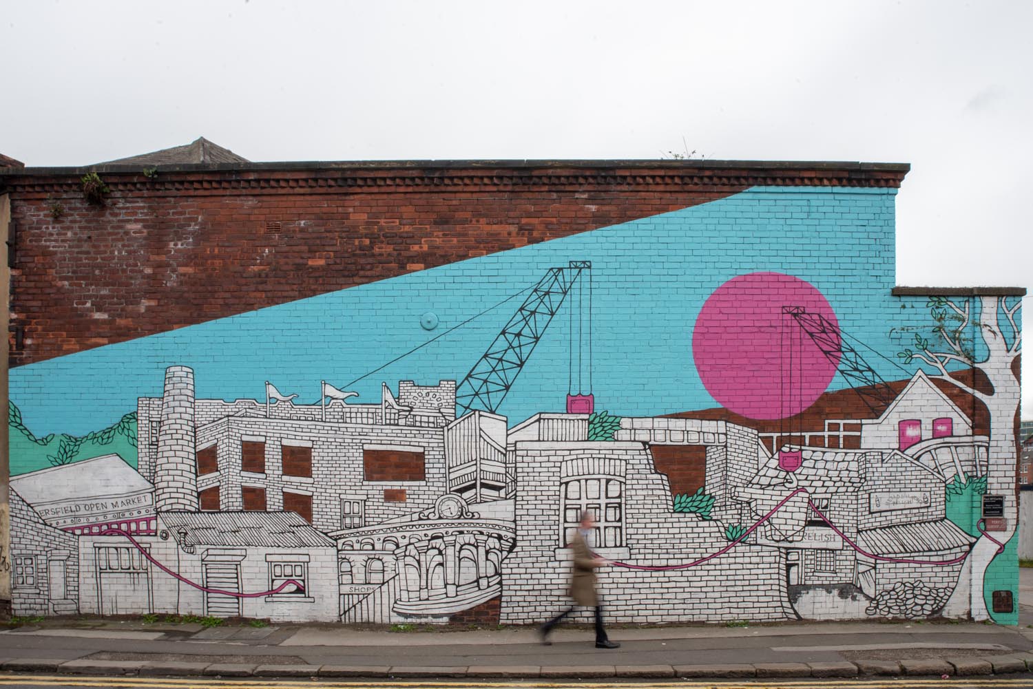 A large mural painted on the side of a brick building depicting an abstract cityscape. The artwork features detailed black-and-white line drawings of buildings, cranes, and architectural elements, accented with bright colors: a turquoise sky, a large pink circle, and pink lines connecting parts of the design. A tree with white branches is included on the right side of the mural. A person is walking along the pavement in front of the artwork.