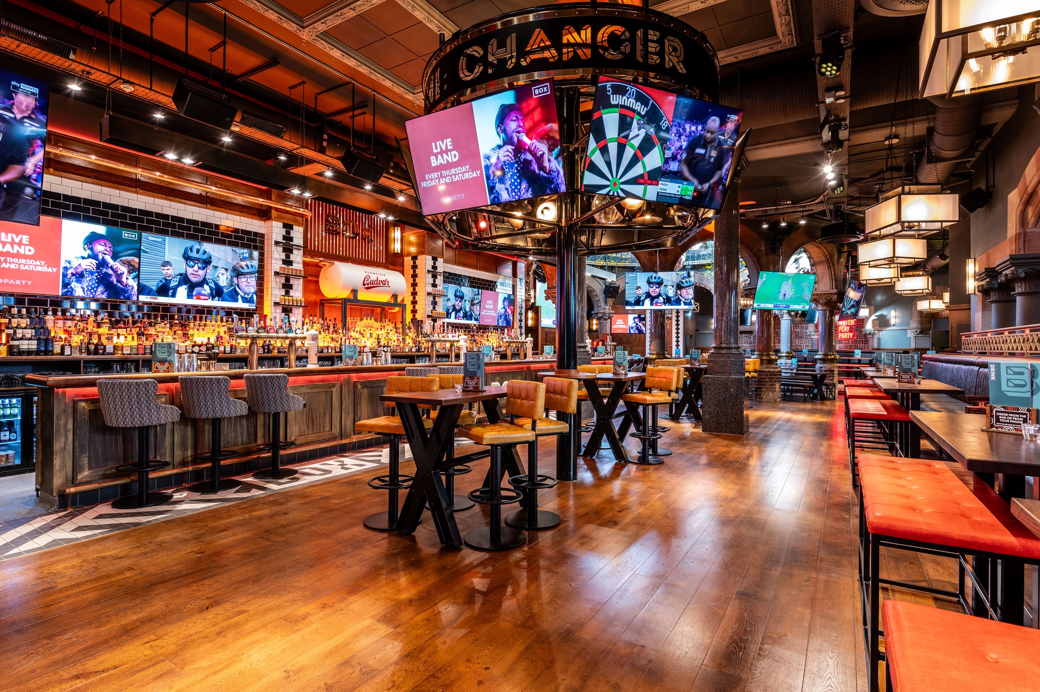 Interior of a modern sports bar with wooden flooring, high tables and stools, and a long bar counter stocked with bottles. Multiple large screens display live sports and event promotions, including a central circular structure with a dartboard and illuminated signage. Warm ambient lighting and industrial-style decor create a vibrant atmosphere.