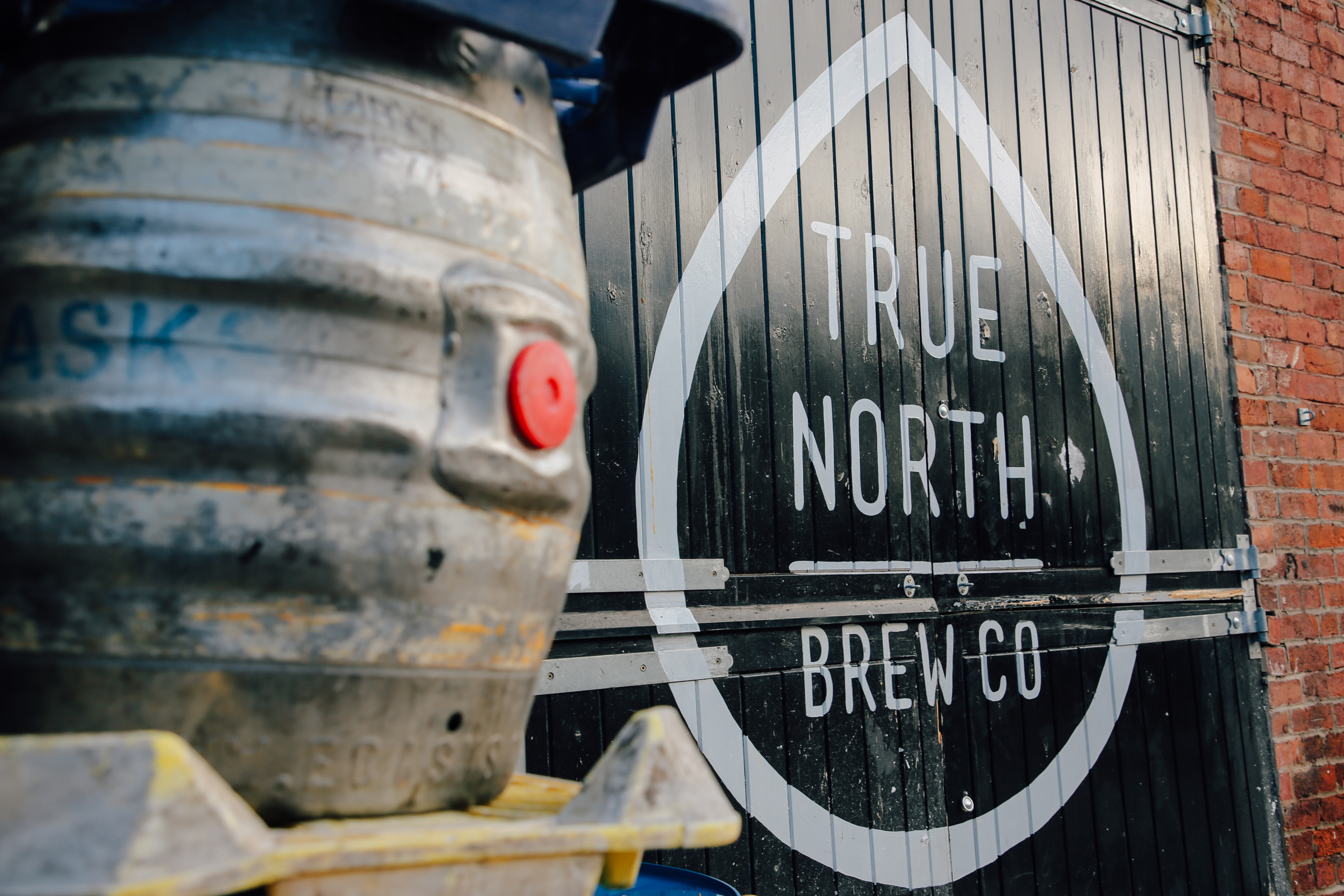 Metal beer keg in the foreground with a black wooden door in the background featuring a large white droplet-shaped outline and the words ‘TRUE NORTH BREW CO’ in bold uppercase letters.