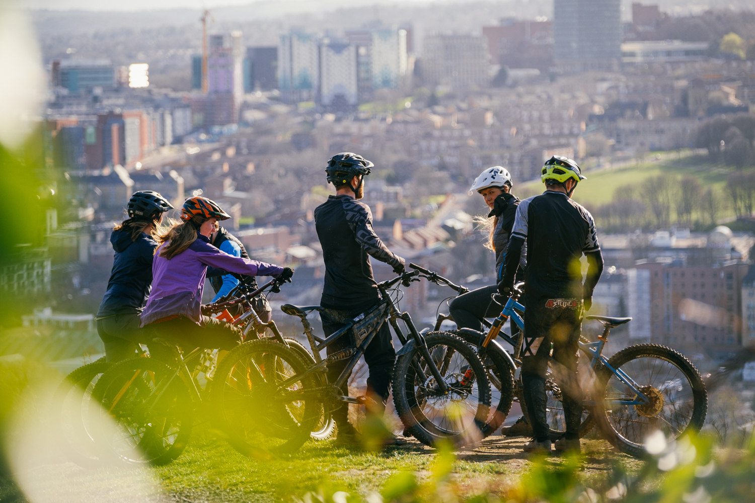 A group of five cyclists wearing helmets and outdoor gear are stopped on a grassy hilltop with mountain bikes, overlooking a cityscape below. The background shows a mix of tall buildings, houses, and green spaces under daylight, with blurred foliage framing the foreground.
