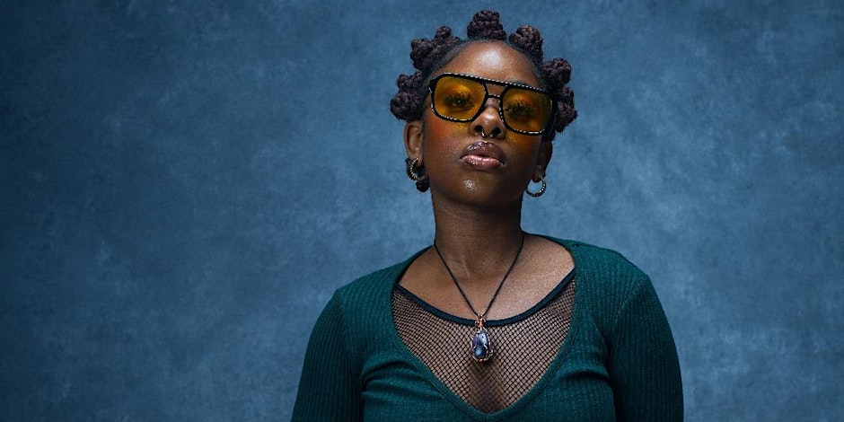 Person wearing a deep green long-sleeve top with a black mesh neckline and a pendant necklace featuring a dark stone, standing against a textured blue background.