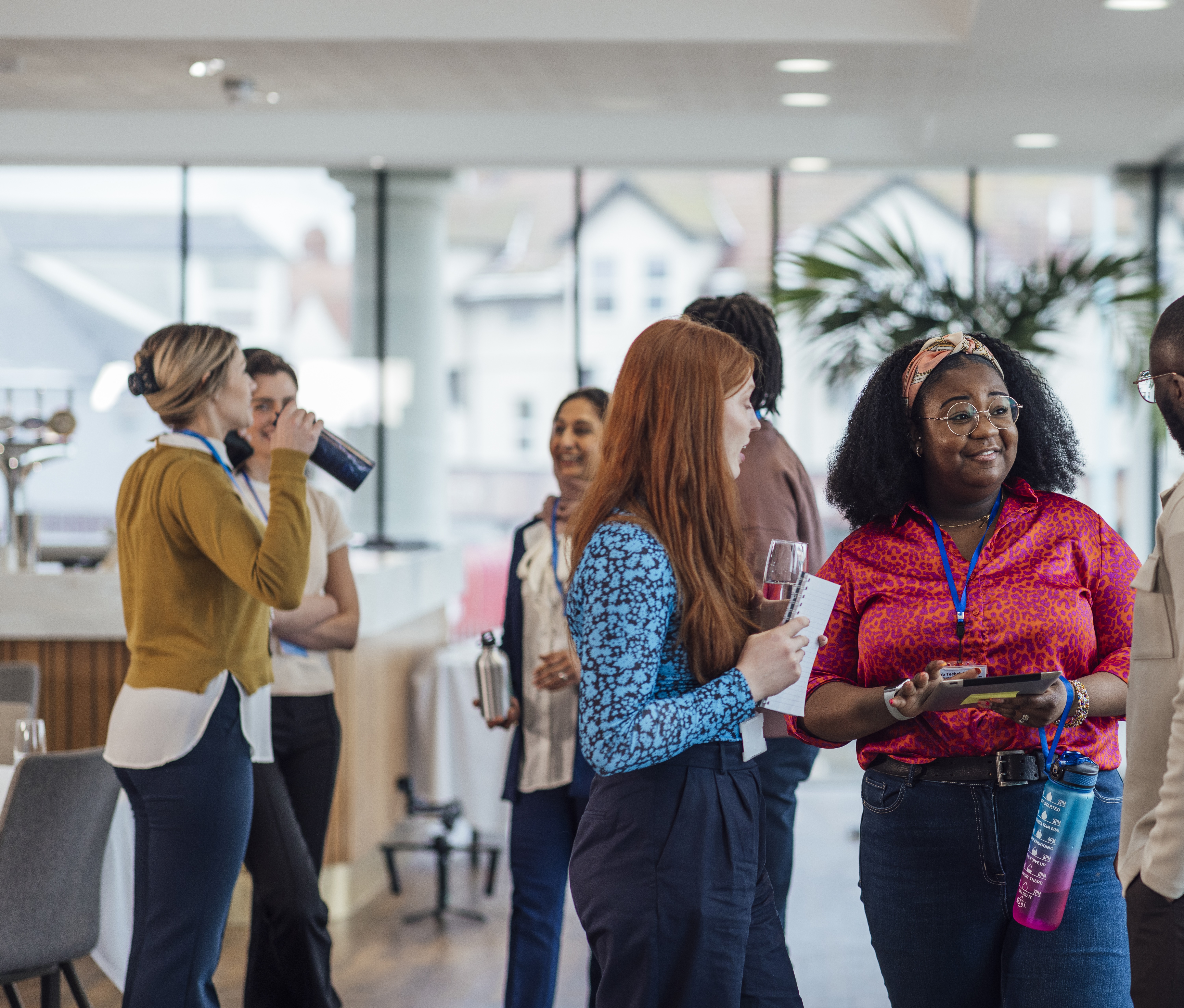 A group of people standing and talking in a modern indoor setting, likely during a networking or social event. The space has large windows with natural light, a bar or refreshment area in the background, and a tall plant adding greenery. Several individuals are holding drinks or notebooks, and one person has a bright pink water bottle. The atmosphere appears casual and professional.