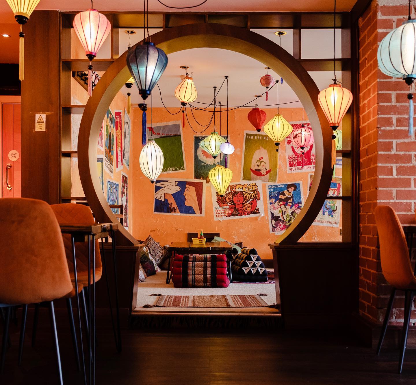 Interior of a cozy restaurant with warm lighting, featuring a circular doorway framing a seating area with low tables and colorful cushions. The walls are decorated with vibrant posters, and multiple hanging lanterns in various shapes and colors create a festive atmosphere.