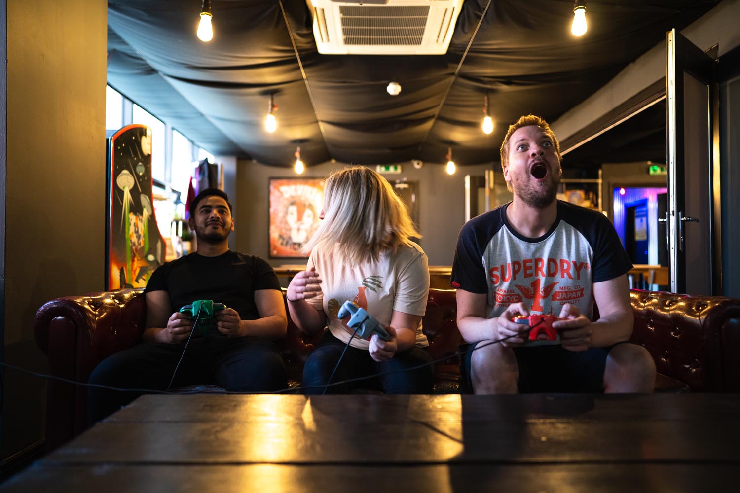 Three people sitting on a leather sofa in a gaming lounge, each holding a retro-style game controller. The room has dim lighting with exposed bulbs, arcade machines in the background, and a black ceiling with visible air conditioning unit.