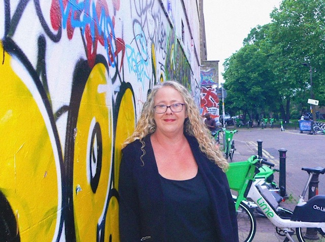 Person with long curly hair wearing a dark jacket, standing next to a wall covered in colorful graffiti featuring large yellow shapes and multicoloured tags. In the background, there are green rental bikes and trees along a street.