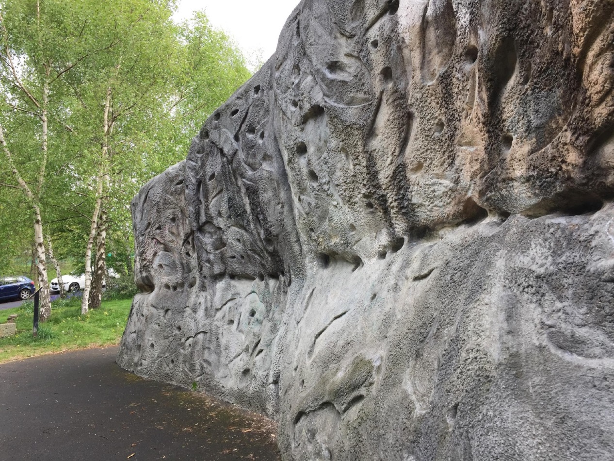 Outdoor climbing wall with various handholds and textures. It is located beside a paved path, surrounded by grass and trees, with parked cars visible in the background.