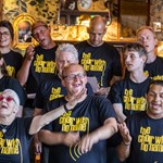 Members of The Choir With No Name singing together in black t-shirts with yellow text, standing in front of a decorative wall with mirrors and lights.