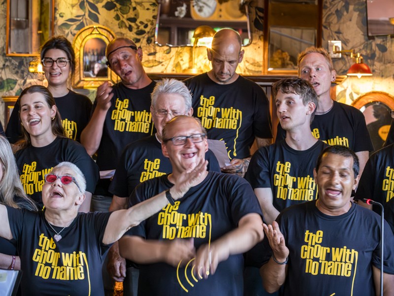 Members of The Choir With No Name singing together in black t-shirts with yellow text, standing in front of a decorative wall with mirrors and lights.