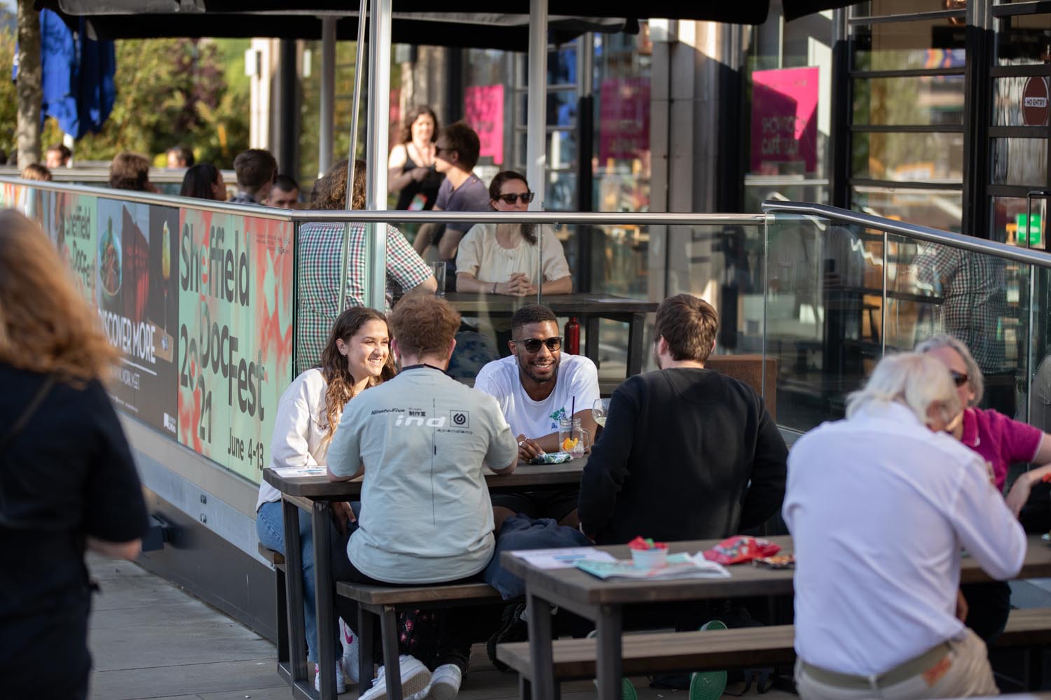 Outdoor seating area of a café or restaurant with several people sitting at wooden tables, eating and drinking. The space is enclosed by glass panels with colorful signage promoting “Sheffield DocFest” and event dates. String lights and umbrellas provide shade, and the background shows more tables and people inside the establishment.