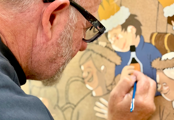 A person is painting a detailed illustration on a large surface. The artwork features cartoon-style characters wearing festive paper crowns, with one character holding a bottle. The painter’s hand is visible holding a fine paintbrush, applying colour to the artwork. The scene suggests a creative process focused on holiday or celebratory imagery.