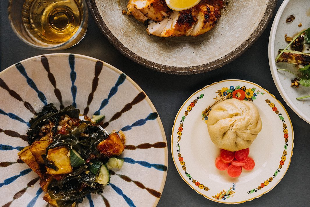 Top-down view of a table with assorted dishes, including a plate of roasted vegetables with greens, a decorative plate holding a steamed bun and pickled radishes, a stone bowl with grilled meat topped with a lemon wedge, and a glass of golden-coloured drink.