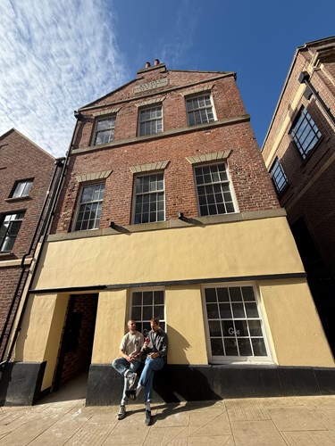 Tall brick building with three stories and a cream-colored ground floor facade. The upper floors have large rectangular windows with stone lintels, and the roofline features a small decorative gable with engraved text. Two people are sitting on the low ledge in front of the building, near a window with the word ‘Güs’ visible on the glass. The scene is lit by bright sunlight under a clear blue sky with wispy clouds.