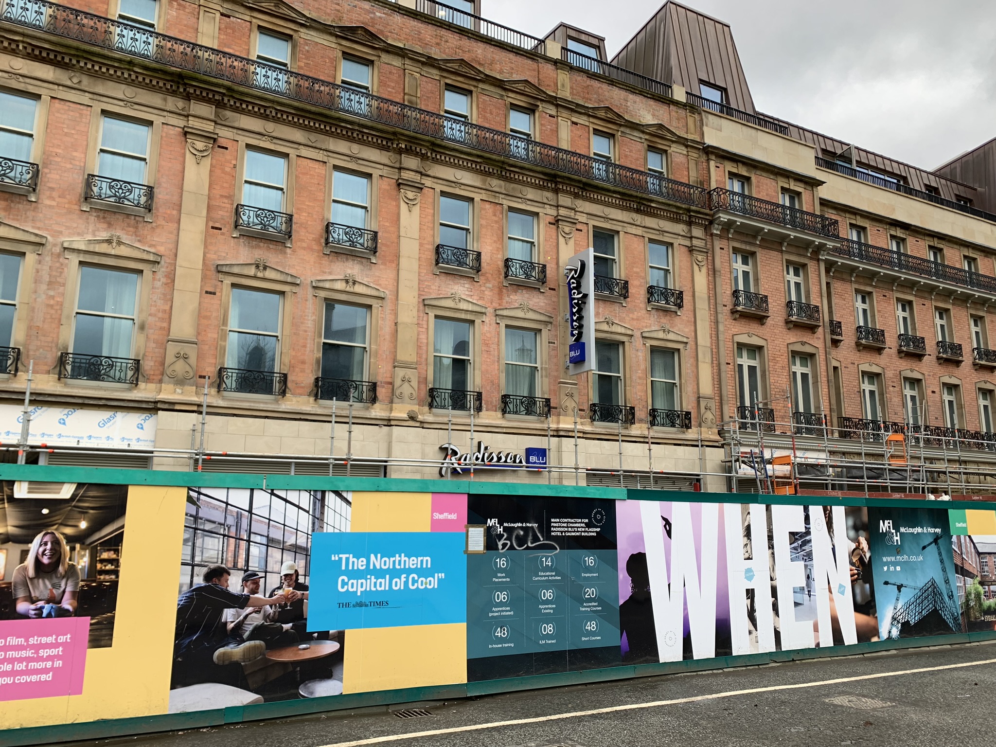 Street view of a historic building with a Radisson Blu hotel sign, featuring ornate windows and black wrought-iron balconies. In front of the building is a colorful construction hoarding with promotional graphics and text, including phrases like “The Northern Capital of Cool” and “WHEN.” The hoarding displays images of people indoors, event details, and branding elements. Scaffolding is visible along the lower part of the building, and the sky is overcast.
