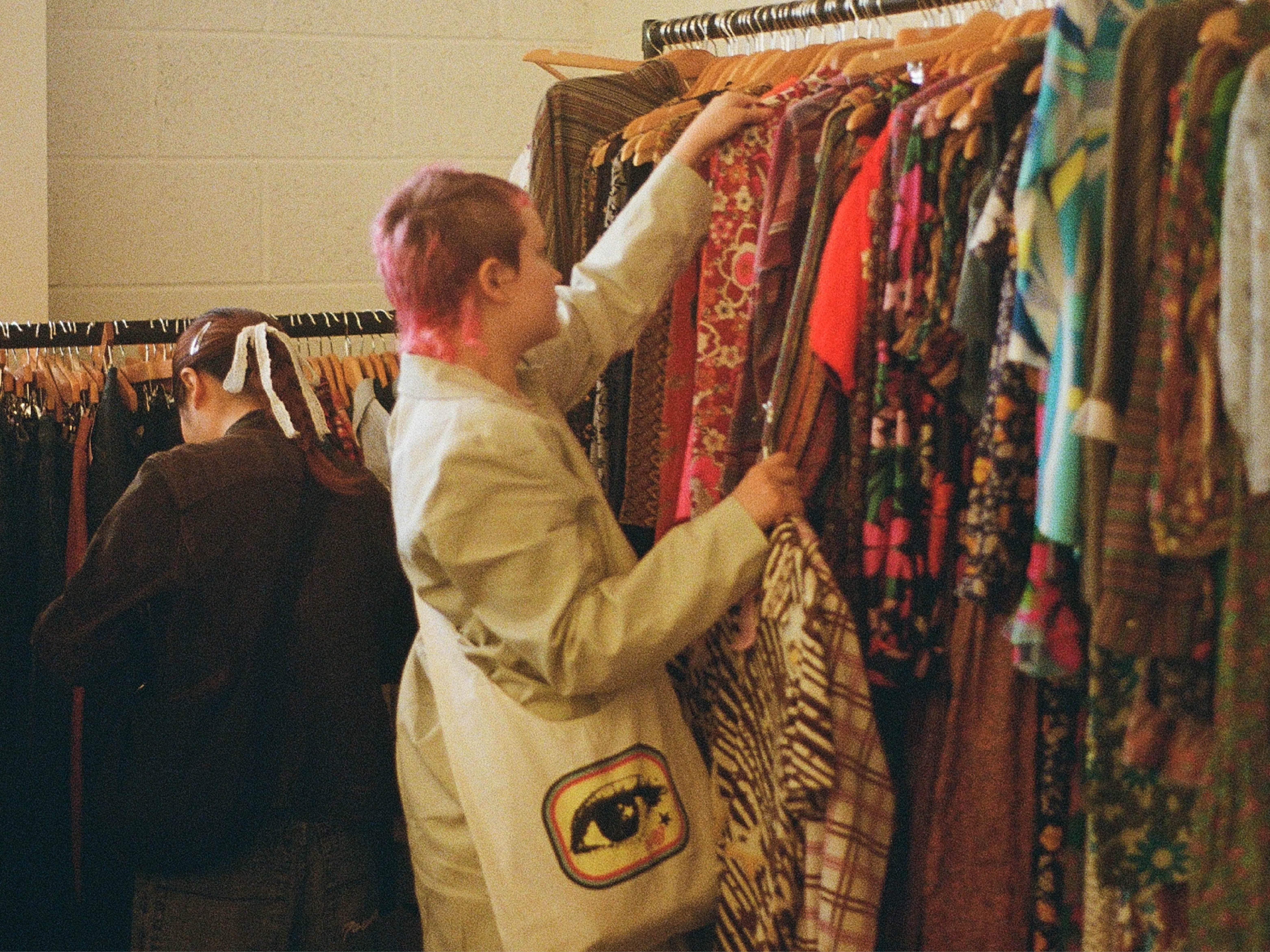 Two people are browsing a clothing rack filled with colorful garments, including patterned dresses and tops. One person with short, pink-tinted hair is wearing a light-colored jacket and carrying a cream tote bag with a bold eye design, while reaching for a piece of clothing. The other person is looking through items on an adjacent rack. The setting appears to be a vintage or second-hand clothing store.